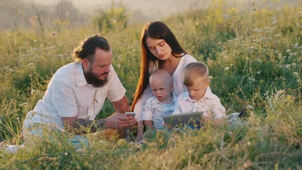 Сім'я з двома дітьми відпочиває на лузі. Грайте зі своїм планшетом і телефоном. Маленькі хлопчики люблять ігри — стокове відео