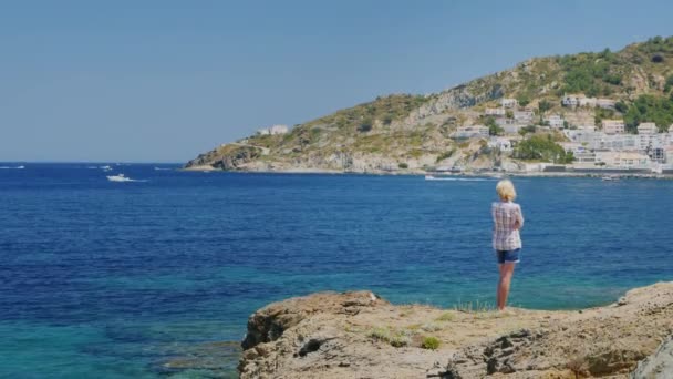 岩の多い海岸で、海を眺めに若い女性が立っています。彼女の前に小さなリゾート スペイン カダケスの町 — ストック動画