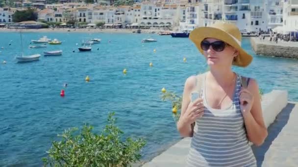 Steadicam shot: Una mujer con sombrero y gafas de sol, caminando por el paseo marítimo. En el contexto de la popular localidad turística de Cadaques en España — Vídeos de Stock