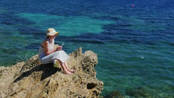 Crane shot: Giovane donna seduta su una roccia, con un cellulare. Affacciato sul bellissimo mare — Video Stock