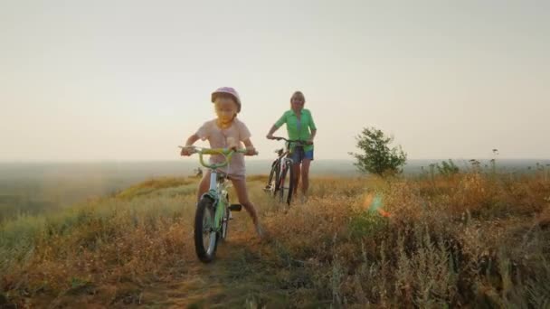 Steadicam shot: Мама учит дочь ездить на велосипеде. Девушка наслаждается солнцем на закате — стоковое видео