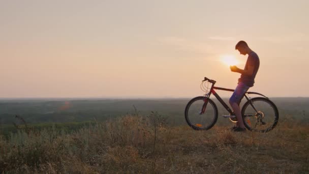 Adolescente sentado en la bicicleta utiliza un teléfono inteligente. En la puesta de sol de su silueta — Vídeos de Stock