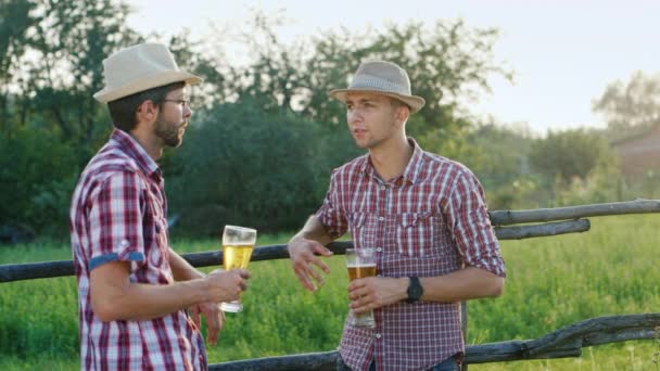 Dois homens rurais com chapéus a beber cerveja numa vedação de madeira. No campo — Vídeo de Stock