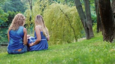 İki arkadaş - genç kadın parkta yeşil çimenlerin üzerinde rahatlatıcı. Tablet keyfini çıkarın. Arkadan Görünüm