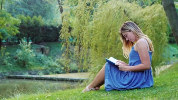 Junge attraktive Frau liest im Park ein Buch und sitzt im Gras — Stockvideo