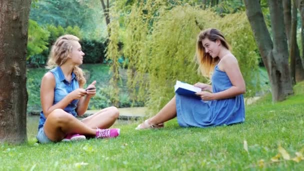 Dos amigas relajándose en el parque - usa el teléfono, lee un libro — Vídeos de Stock