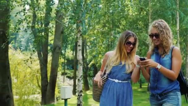 Steadicam shot: Dos amigas pasan por el parque, disfrutan de un smartphone, sonriendo — Vídeos de Stock