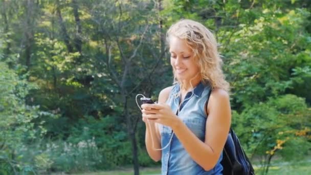 Ung kvinna promenader i parken, lyssna på musik i hörlurar — Stockvideo