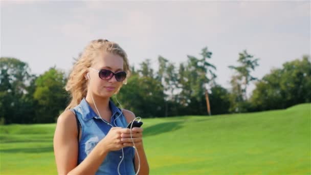 Giovane donna si avvicina a un grande campo da golf, ascoltando musica sulle cuffie in mano tenendo lo smartphone — Video Stock