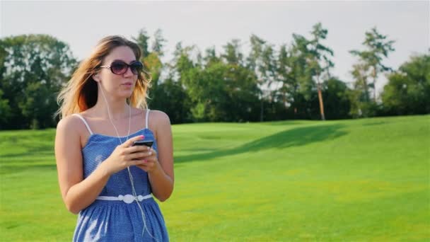 Steadicam shot: jonge vrouw gaat in de buurt van een grote golfbaan, luisteren naar muziek op de hoofdtelefoon in handen met slimme telefoon — Stockvideo