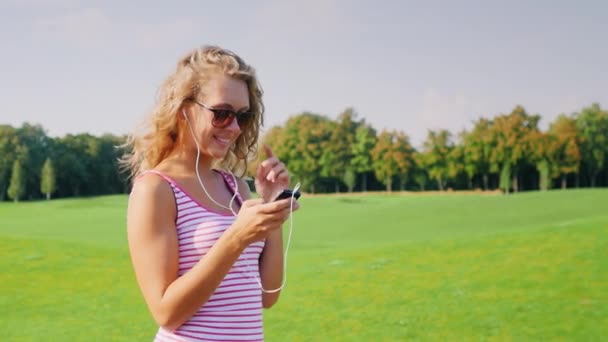 Jovem mulher se aproxima de um grande campo de golfe, ouvindo música em fones de ouvido em mãos segurando telefone inteligente — Vídeo de Stock