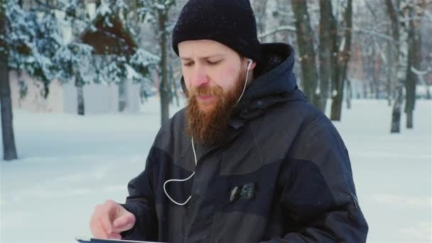 Steadicam geschoten: aantrekkelijke bebaarde man wandelen in een winter park, geniet van een tablet — Stockvideo