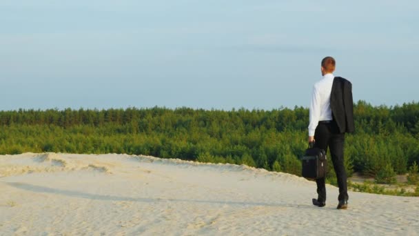 Un hombre de negocios despreocupado caminando sobre la arena, lleva una chaqueta sobre su hombro. Vista trasera — Vídeos de Stock