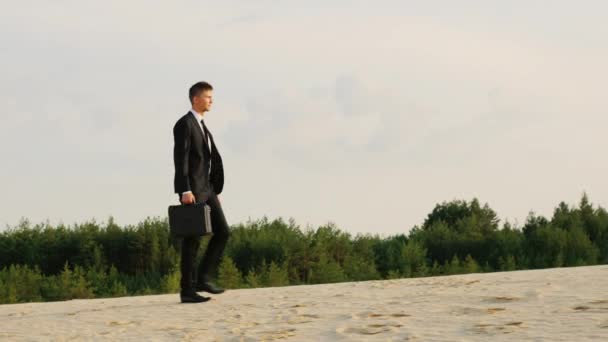 Joven hombre de negocios con maletín caminando sobre la arena en un bosque. Lejos de preocupaciones — Vídeo de stock