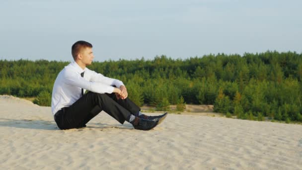 Crane shot: Un joven en un traje de negocios sentado en la arena y admirando el paisaje del bosque. El concepto - el descanso del trabajo — Vídeos de Stock