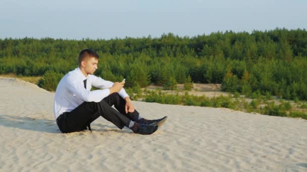 Zawsze w kontakcie. Młody biznesmen z telefonu siedzi na piasku na tle malowniczego lasu — Wideo stockowe