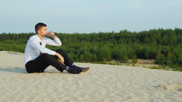Молодой бизнесмен пьет алкоголь из фляжек, сидя на песке. Концепция - проблемы, банкротство, бизнес, крах — стоковое видео
