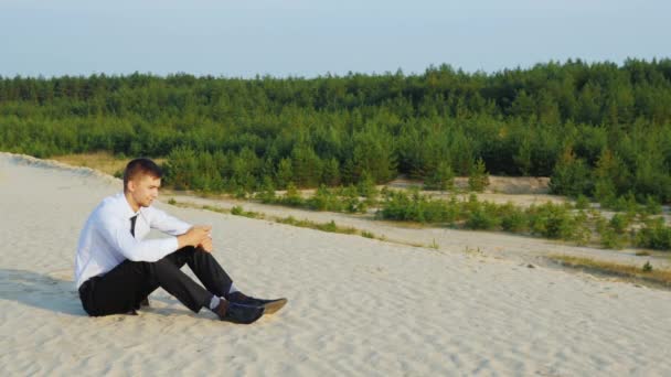 Joven hombre de negocios escribiendo un mensaje en su teléfono, sentado en la arena contra el telón de fondo de un pintoresco bosque. Concepto - siempre conectado — Vídeos de Stock