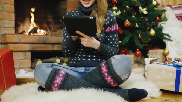 Joven hermosa mujer sentada junto a la chimenea, disfrutar de la tableta. Decoraciones de Navidad detrás de ella, calcetines para regalos — Vídeo de stock