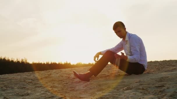 Giovane uomo d'affari premuroso seduto sulla sabbia, guardando la rottura delle mani della sabbia al tramonto. Concetto: la transitorietà del tempo, il cambiamento — Video Stock