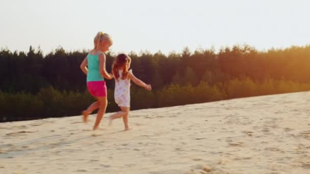 Steadicam toma de cámara lenta: Dos chicas alegres corriendo una carrera en la arena al atardecer. Juegos para niños, una infancia feliz — Vídeo de stock