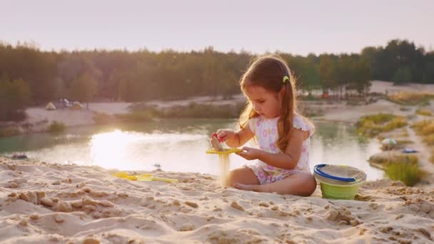 Een meisje van drie jaar spelen in het zand op een achtergrond van meer en recreatieve — Stockvideo