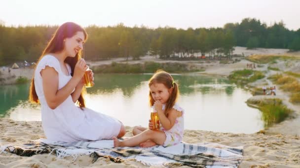 Mamá con una hija tres años relajándose en la playa junto al lago, beber jugo de botellas con tubos, riendo — Vídeos de Stock