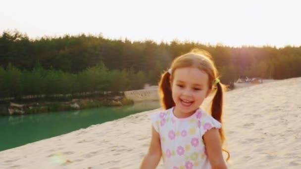 Steadicam slow motion shot: ξέγνοιαστες χαρούμενο κορίτσι τριών ετών που τρέχουν στην άμμο. Με φόντο τις πανέμορφες λίμνες και τα δάση. — Αρχείο Βίντεο