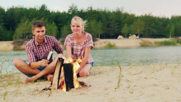 Przyjaciele mężczyzna i kobieta siedzą wokół ogniska, porcję pieczeni na kije. — Wideo stockowe