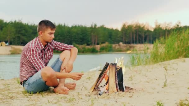 Young attractive man sitting by the fire alone. Fries marshmallow on a stick. Against the background of the forest and the river — Stock Video
