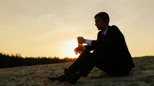 Ένας νεαρός άνδρας σε ένα επαγγελματικό κοστούμι κάθεται στην άμμο και πίνοντας μπύρα από ένα μπουκάλι. Έννοια - απογοητεύσεις, πτώχευση, κατάθλιψη — Αρχείο Βίντεο