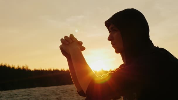 Ein junger Mann mit Kapuze sitzt bei Sonnenuntergang, es ist nachdenklich, träumend — Stockvideo