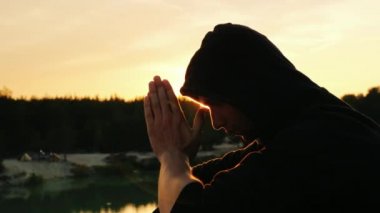 Gün batımında dua genç adam. Kamera hood için profilde oturuyor ellerini sıktı birlikte dua