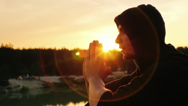 Egy fiatal férfi, egy kapucnis súgta az imádság a naplemente. Ül a profil — Stock videók