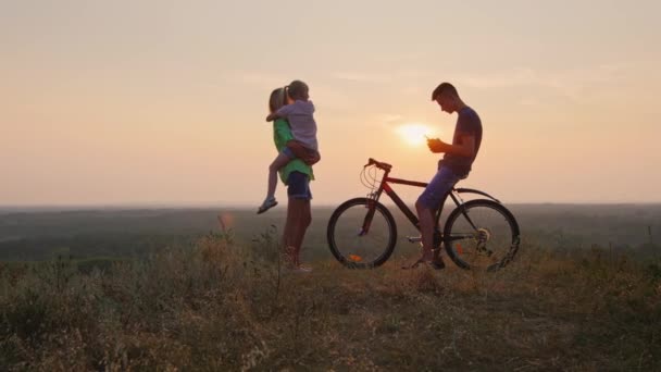 La famille accueille le coucher du soleil. Fils est assis sur le vélo et est le téléphone, mère et fille regardant le soleil couchant — Video