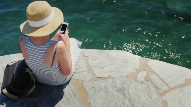 Турист в шляпе сидит на набережной, используя мобильный телефон. Камера движется к заливу с яхтами — стоковое видео