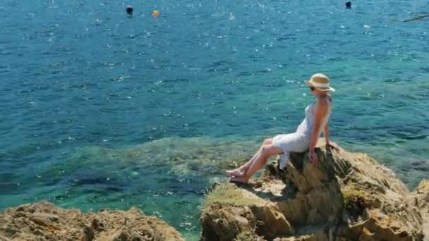 Женщина-туристка в шляпе сидит на скале и восхищается морем — стоковое видео