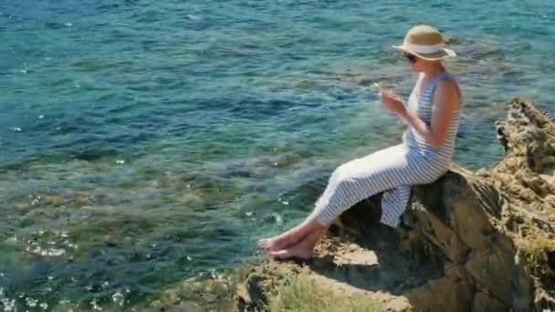 Steadicam shot: Mujer joven sentada en una roca, usando un teléfono móvil. Con vistas al hermoso mar — Vídeo de stock