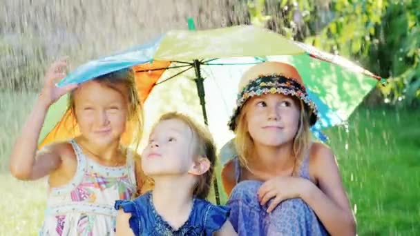 Tres alegre chica despreocupada bajo el paraguas de los colores del arco iris. Hay una cálida lluvia. Feliz infancia. — Vídeos de Stock