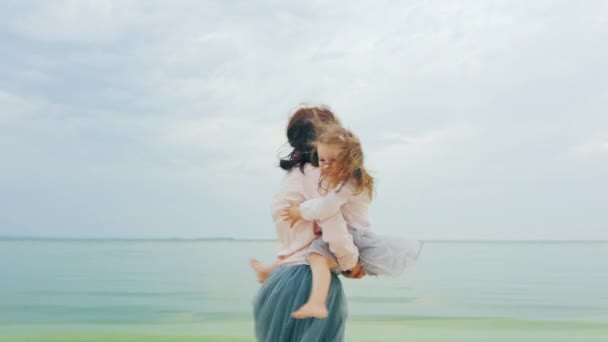 Mamãe brinca com sua filha, circulando-a, tanto rindo quanto se divertindo. Contra o fundo do mar e do céu — Vídeo de Stock