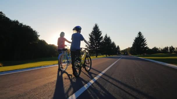 Σταθερα shot: γυναίκα και γιος που περπατούν στο πάρκο με ποδήλατα. Στο ηλιοβασίλεμα. Πίσω όψη — Αρχείο Βίντεο