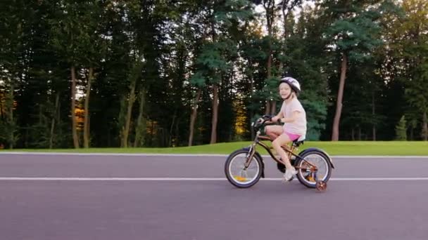 Menina de cinco anos em um capacete andando de bicicleta. No parque bem guardado em um contexto de árvores — Vídeo de Stock