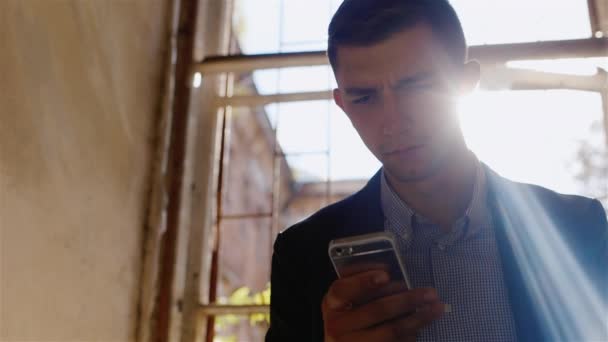 Joven empresaria infeliz escribiendo sms. Sentado en una casa abandonada cerca de la ventana. Los rayos del sol brillan por detrás — Vídeo de stock