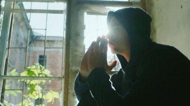 Kapüşonlu genç bir pencere eşik üzerinde oturur, dua etmek için başını ellerini tutarak. Güneş ışınlarında
