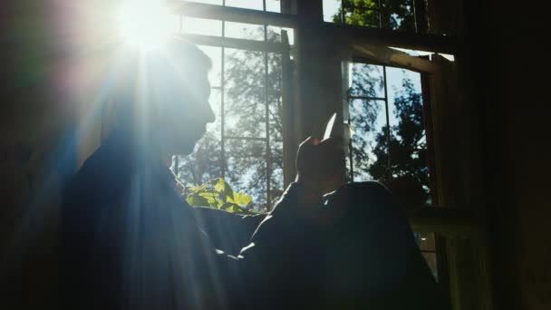 El joven empresario en una casa abandonada. Se sienta en el alféizar de la ventana, usa un teléfono. Silueta al sol — Vídeo de stock