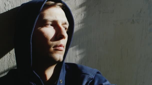 Upprörd ung Hooded man sitter nära fönstret. Solen är lysande fläckar på hans ansikte — Stockvideo
