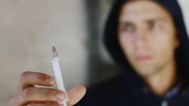 Kapüşonlu genç bir adam uyuşturucuyla şırıngaya bakıyor. Kavram: suç, uyuşturucu bağımlılığı