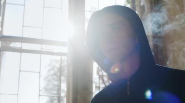Genç terk edilmiş bir evde sigara içiyor. Güneşin arkasında parlıyor, hepsi sigara sisinde.