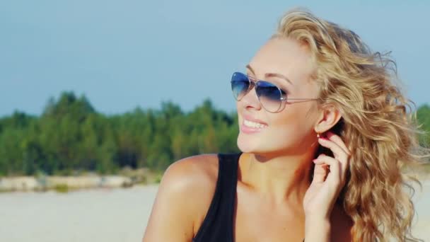 Mladá žena ve slunečních brýlích se usmívá do kamery. Vítr si hraje s jejími vlasy — Stock video