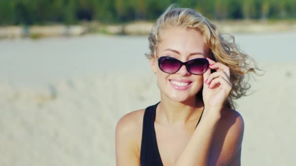 Красива молода жінка посміхається в камеру, дивлячись на сонцезахисні окуляри. На пляжі в сонячний день — стокове відео